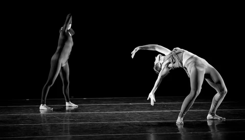 Two dancers in dramatic poses on a dark stage, captured in high-contrast black and white lighting.