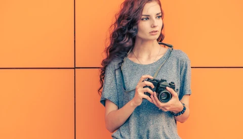 Woman with long wavy hair holding a vintage film camera against an orange background.
