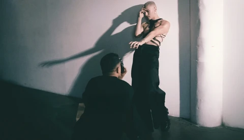 Shirtless man against white wall with dramatic shadow of another person cast across his torso.