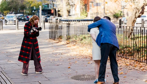 Man in blue blazer posing while woman in red plaid coat photographs him on an urban sidewalk.