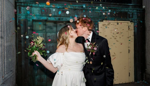 Newlyweds kissing in front of a teal doorway decorated with hanging paper lanterns and confetti.
