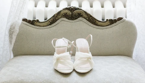 Pair of white infant shoes displayed on a cream-colored vintage chair with ornate wooden frame.