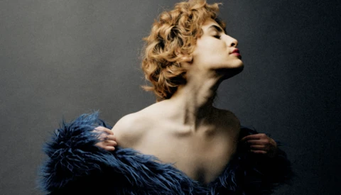 Woman with curled blonde hair in profile, shoulders bare, wearing dark blue feathered fabric.