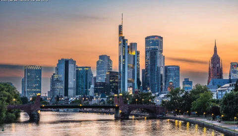 Frankfurt skyline at sunset reflected in the Main River, with illuminated skyscrapers against an orange and pink sky.