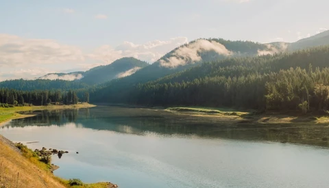Alpine lake with forested mountains reflected in still water under partly cloudy sky.