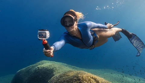 Snorkeler swimming horizontally above a large underwater boulder while holding an action camera.