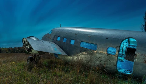 Abandoned aircraft fuselage with detached wing panel sits in overgrown field under dramatic blue sky.