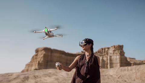 Operator wearing VR headset controls a small quadcopter drone in flight above desert terrain.