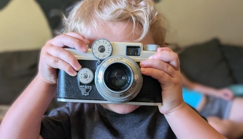 Young child holding a vintage rangefinder camera up to their face, obscuring their head.