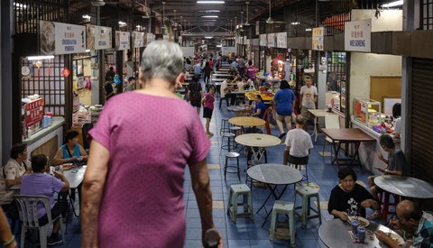 Woman in purple shirt walking through crowded indoor market or fair with vendor booths and tables.