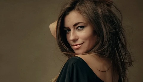 Woman with long dark hair looking over shoulder against neutral tan background.