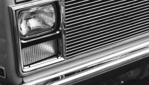 Close-up black and white photograph of a classic car's front end, showing rectangular headlight and horizontal grille detail.
