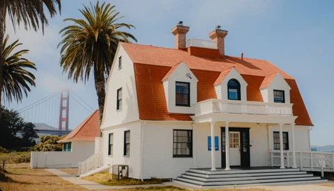 White Victorian-style house with distinctive orange-red mansard roof and dormer windows, palm trees nearby, suspension bridge visible in background.