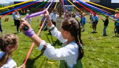 Children holding colored ribbons around a maypole on a grass field during a community event.