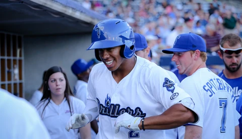 Baseball player in blue uniform smiling while being congratulated by teammates in dugout.