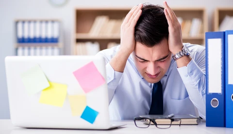 Stressed man at desk with laptop covered in sticky notes, holding his head in frustration.