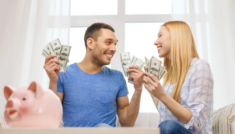 Couple holding cash and smiling at each other indoors by a window.