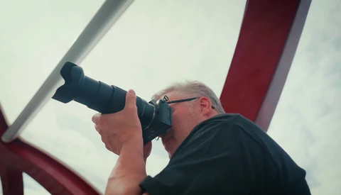 Photographer holding telephoto lens up to eye while shooting outdoors against cloudy sky.