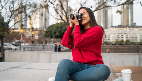 Woman in red jacket holding binoculars to her eyes while sitting outdoors near urban architecture.