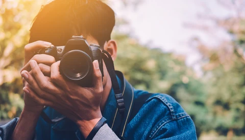 Photographer holding a DSLR camera up to their face, composing a shot outdoors in natural sunlight.