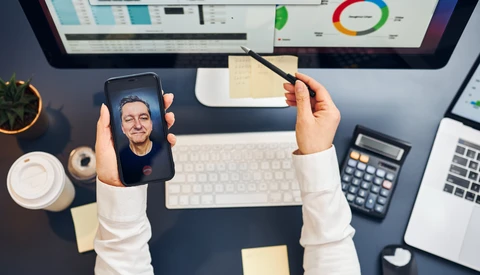 Person holding smartphone displaying a portrait photo while working at a desk with monitor and analytics dashboard.