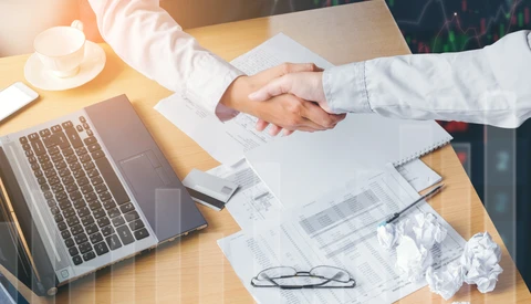 Two people in white shirts shake hands over a desk during a business meeting.