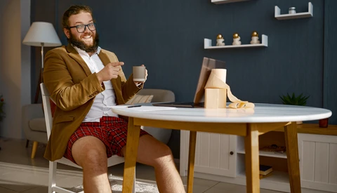 Bearded man in mustard blazer and plaid shorts sits at round table with laptop and coffee cup.