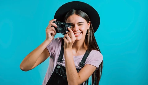 Young woman holding vintage film camera up to her face, wearing black hat and overalls against bright turquoise background.