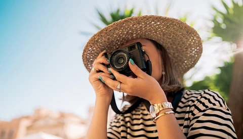 Woman in striped shirt and straw hat holding vintage camera to her face.