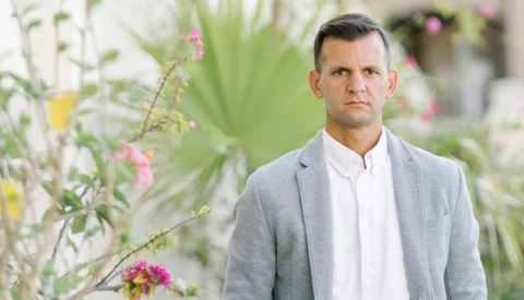 Man in light gray blazer and white shirt standing in front of blooming flowers and foliage.