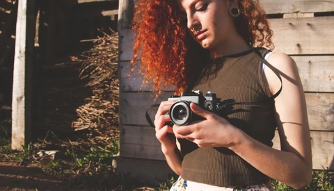 Woman with red curly hair holding vintage film camera outdoors.