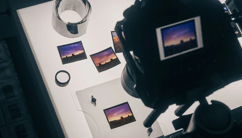 Overhead view of a photographer's workspace with printed sunset landscape photos, a camera, glass of water, and lens cap on white surface.