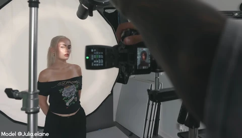 Model posing during studio photoshoot with professional cinema camera and large softbox lighting setup.