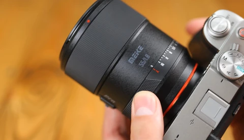Hand holding a mirrorless camera with a black telephoto lens attached against a warm, blurred orange background.
