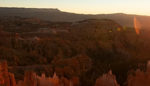 Golden sunrise light illuminates layered hoodoos and forested ridges in a vast canyon landscape.