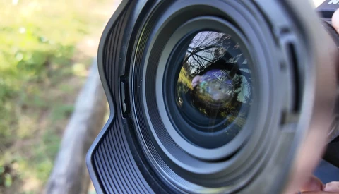 Close-up of a camera lens front element with shallow depth of field, showing tree branches and landscape reflected in the glass.