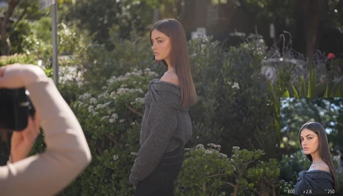Pregnant woman in dark sweater posing among garden foliage while photographer frames shot in foreground.