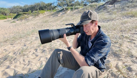 Photographer seated on sand using a telephoto lens while shooting at a beach location.