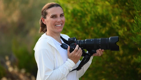 Photographer holding a professional camera with telephoto lens, smiling at camera against blurred green foliage background.