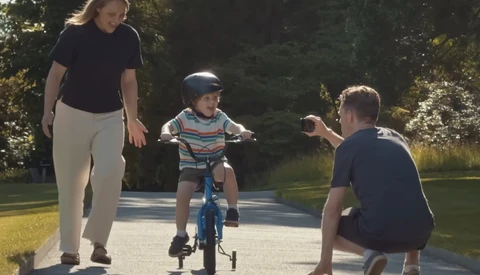 Woman supervises young child riding blue bicycle with training wheels on a paved path as man crouches nearby.