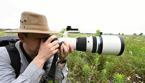 Photographer holding a white telephoto lens while looking through a camera in an overgrown field.