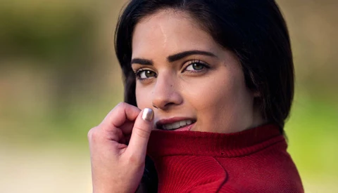 Woman in red sweater with hand near face, outdoor portrait with blurred green background.