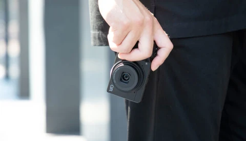 Person in dark clothing holding a compact action camera with a circular lens against their body.