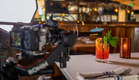 Professional video camera on tripod filming colorful cocktails on a white table in a bar setting.