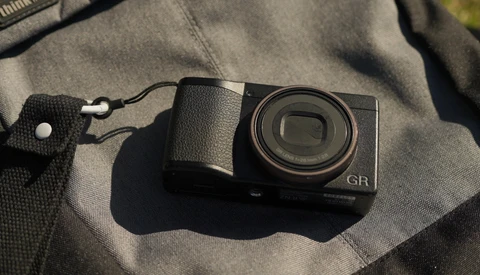 Ricoh GR compact digital camera with textured grip sitting on a gray fabric surface with a wrist strap attached.