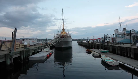 Large cargo ship docked at industrial waterfront with smaller boats and wooden pilings in foreground.