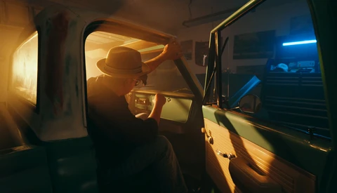 Silhouetted figure wearing a hat inside a vehicle at night, illuminated by warm and cool ambient light.