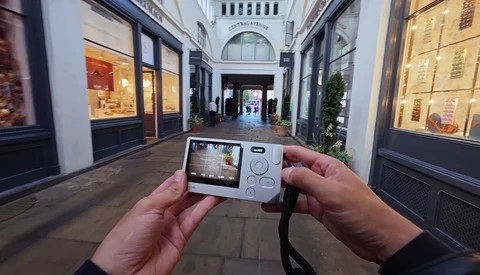 Hands holding a compact digital camera displaying an arcade game on its LCD screen in an upscale shopping passage.