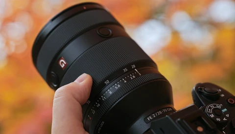 Hand holding a telephoto lens with focus ring and depth-of-field scale visible against blurred autumn foliage background.