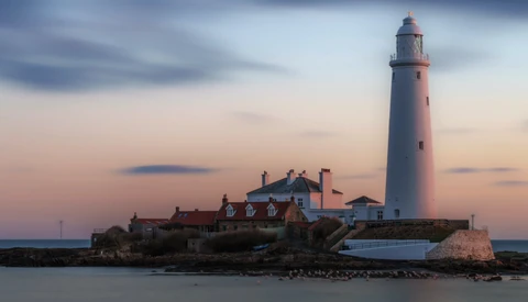 Lighthouse with attached keeper's house on rocky outcrop at twilight, warm sky with cool water below.